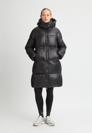 Zwarte pufferjas met een hoge kraag, voorzien van een glanzende buitenzijde, ritsluiting en twee zijzakken; gedragen over zwarte leggings en sneakers.