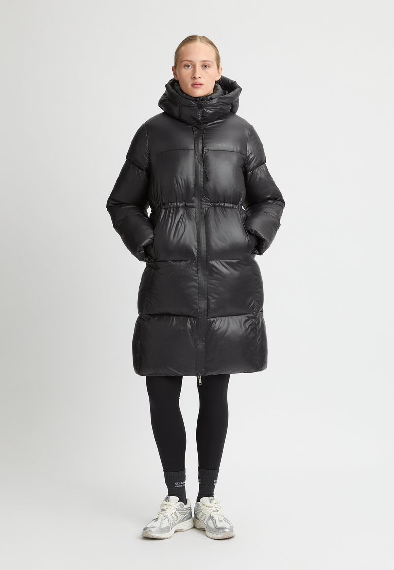 Zwarte pufferjas met een hoge kraag, voorzien van een glanzende buitenzijde, ritsluiting en twee zijzakken; gedragen over zwarte leggings en sneakers.
