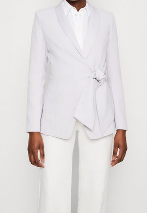 Kvinde iført en lys lavendel blazer med en sidelås over en hvid skjorte med knapper og hvide bukser mod en ensfarvet baggrund.