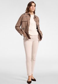 Camisa marrón de botones con bordado floral, usada sobre un suéter de punto trenzado color crema, combinada con pantalones beige claros y zapatos planos oscuros.