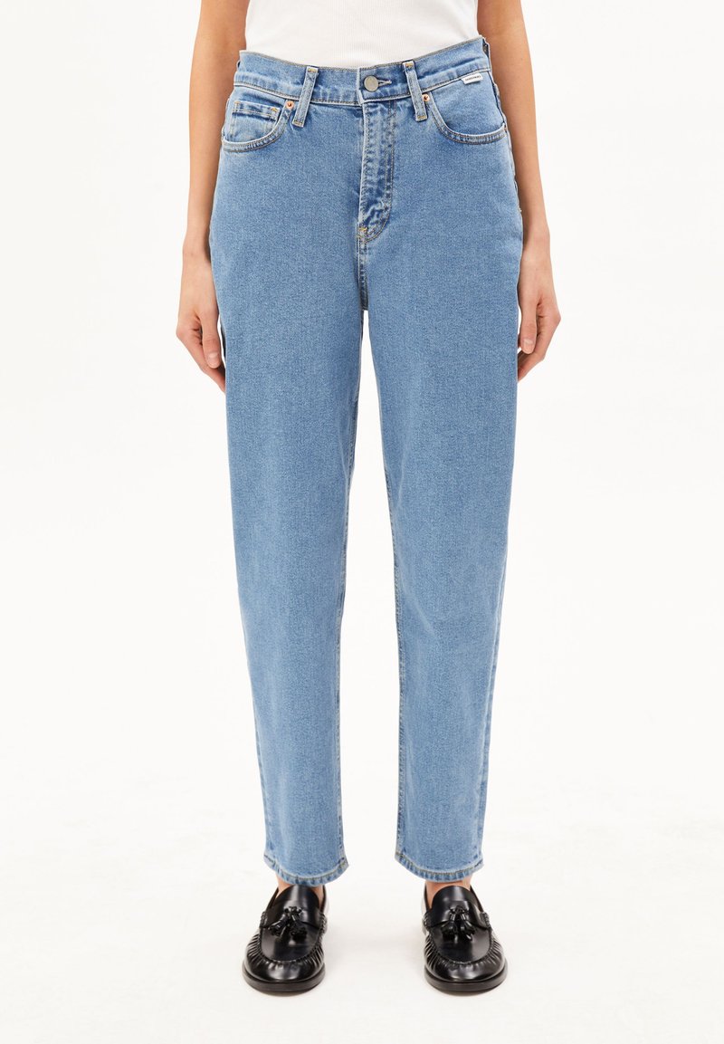 Unterkörper einer Person, die hellblaue, hoch taillierte, gerade geschnittene Jeans und schwarze Tasselloafer trägt und vor einem einfarbigen Hintergrund steht.