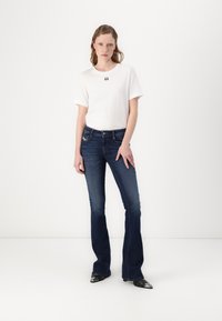 Weißes kurzärmeliges T-Shirt mit einem schwarzen Graphic, kombiniert mit dunkelblauen Schlagjeans. Die Jeans verfügen über einen Knopfverschluss und Taschen.