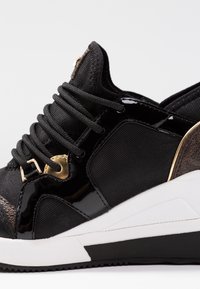 Baskets noires avec une tige en tissu texturé, des accents en vernis brillant et des éléments dorés. Dotées de lacets noirs épais et d'une semelle blanche.