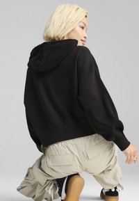 Felpa nera corta con cappuccio regolabile e maniche voluminose, abbinata a pantaloni beige chiaro ampi con tasche sul retro.