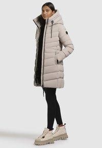Manteau rembourré beige clair avec capuche, fermeture éclair à l'avant, coutures verticales et poches latérales, associé à des bottines blanches à la cheville et à des leggings noirs.