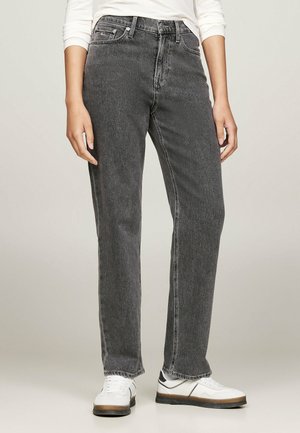 Personne portant un jean noir délavé à coupe droite, des baskets blanches avec des semelles en caoutchouc, et un haut à manches longues blanc, debout sur un fond uni.