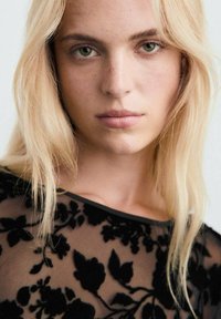 Črn prosojen top z cvetličnimi vzorci, teksturiranimi detajli in širokim izrezom. Model ima dolge blond lase in subtilno ličenje.