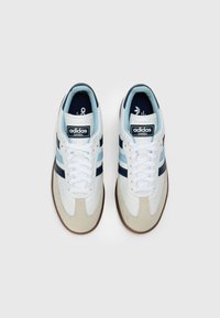 White leather sneakers with blue accents, featuring a suede toe cap and three navy stripes. Classic design with a flat rubber sole.