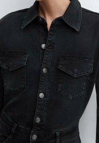 Camisa de mezclilla negra con cierre de botón, cuello clásico, dos bolsillos frontales y adornos en plata. Tejido texturizado con un acabado pulido.