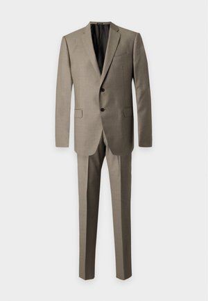 Homme portant un costume marron clair avec une chemise blanche et des chaussures habillées noires, debout avec une main dans la poche sur un fond uni.