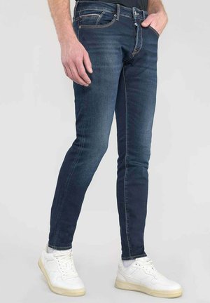 Jean slim-fit bleu foncé en denim, avec un design standard à cinq poches et des coutures contrastées, associés à des sneakers blanches.