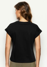 Sort kortærmet t-shirt med rund halsudskæring. Glat tekstur, tætsiddende design og opfoldede ærmer. Baggrunden er neutral.