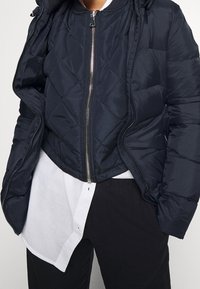 Námořnická prošívaná pufrová bunda se zipem vpředu, žebrovaným límcem a kontrastní podšívkou, přeložená přes bílou košili a černé kalhoty.