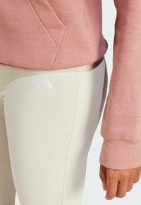 Svetlo roza dukserica s žepom in rebrastimi manšetami, kombinirana s smetanastimi leginami, ki imajo bel logo na stegnu.