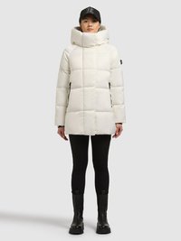 Fehér steppelt dzseki magas gallérral és cipzáras zsebekkel, fekete leggingsszel és masszív fekete csizmával párosítva, fekete sapkával viselve.