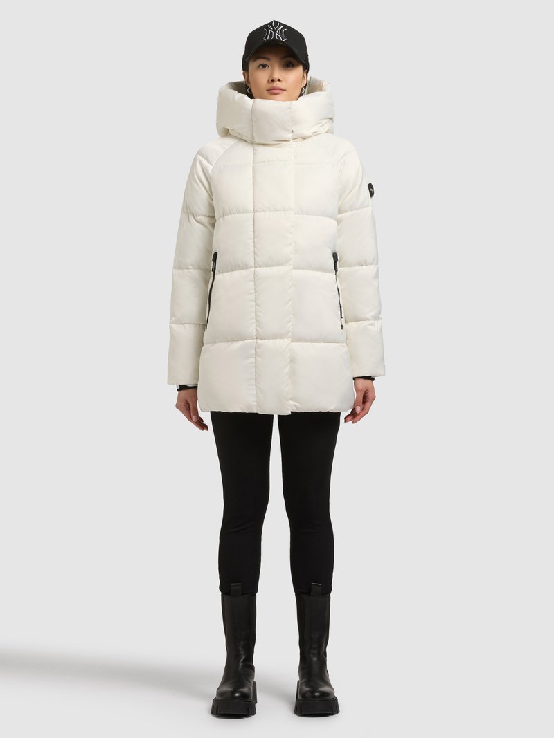 Fehér steppelt dzseki magas gallérral és cipzáras zsebekkel, fekete leggingsszel és masszív fekete csizmával párosítva, fekete sapkával viselve.