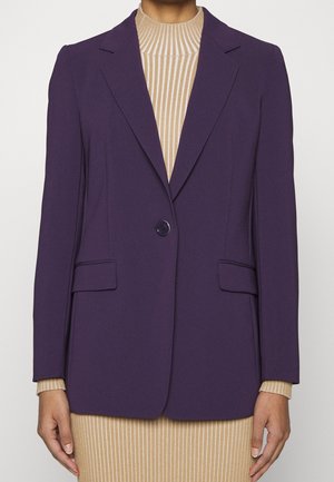 Person iført en skræddersyet lilla blazer med én knap over en beige ribstrikket rullekræls kjole, stående med afslappede hænder langs siderne.