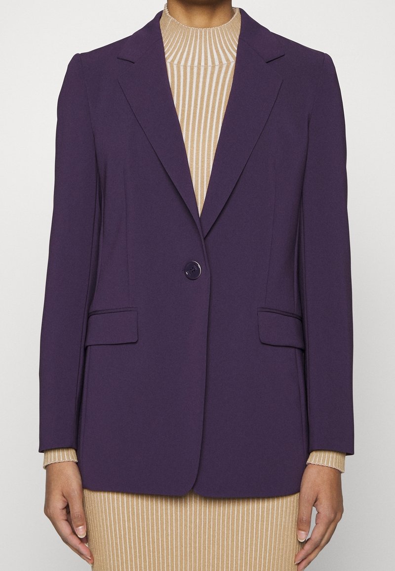 Personne portant un blazer violet ajusté avec un bouton sur une robe beige à col roulé côtelée, debout les mains détendues le long du corps.