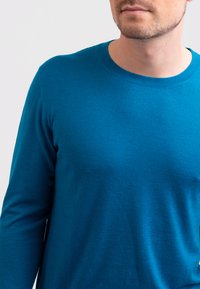 Tealstickad tröja med rund halsringning och långa ärmar. Slät textur utan synliga mönster eller detaljer. Närbild.