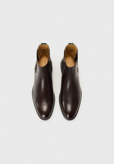 Botas de tobillo de cuero marrón con un diseño elegante, textura suave, puntera afilada y una pestaña verde en el talón. El interior cuenta con un forro beige.