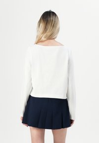 Žena s dlouhými blond vlasy, oblečená v bílé dlouhé rukávové blůze a tmavě námořnické plisované mini sukni, stojí zády.