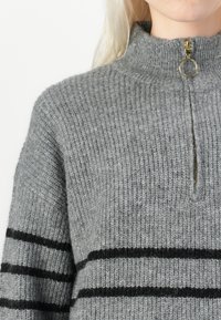 Pull en tricot gris avec un col montant et une fermeture éclair, présentant des rayures horizontales noires et une texture côtelée sur l'ensemble.