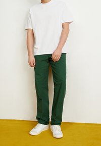 White cotton t-shirt paired with green trousers, featuring a straight cut. Worn with white sneakers on a mustard yellow carpet background.