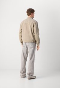 Chaqueta de pana beige con cuello, puños abotonados y textura suave, combinada con jeans gris claro y zapatillas marrones. Vista trasera mostrada.