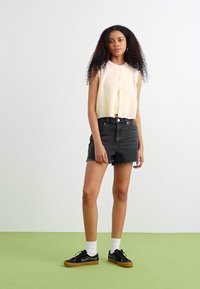 Cardigan senza maniche di colore crema con bottoni, abbinato a pantaloni corti in denim neri a vita alta e scarpe da ginnastica nere con suola beige.