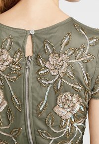 Olijfgroene stoffen top met ingewikkelde bloemengestikte in metallic pailletten, sleutelgatopening aan de achterkant met ritsluiting, en gedetailleerde bladvormen.