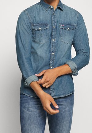 Mann krempelt die Ärmel eines blauen Jeanshemds mit zwei Brusttaschen und passenden blauen Jeans hoch.