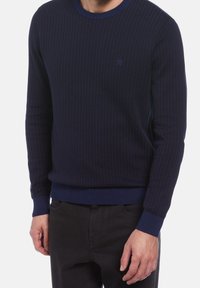 Pull en maille bleu marine avec une texture côtelée, un col rond et des poignets élastiques. Dispose d'un petit logo brodé sur la poitrine.