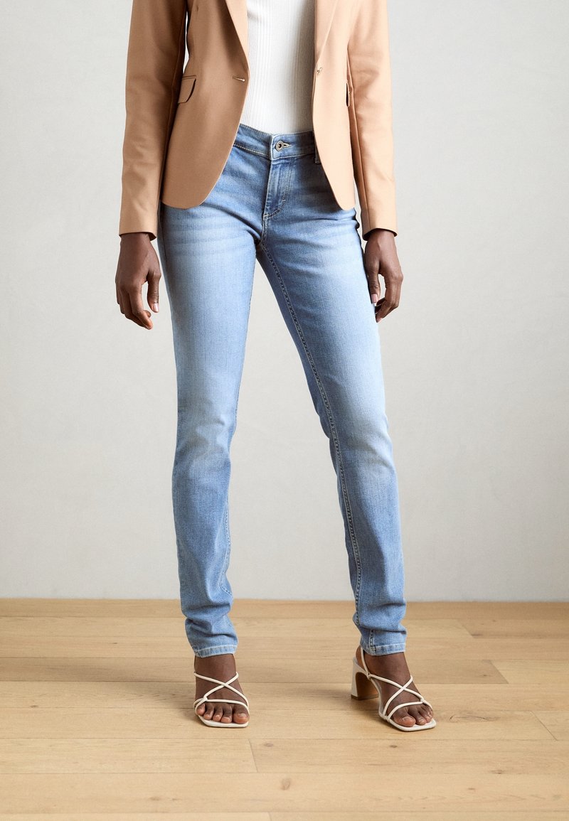 Ljusblå jeans i slim fit, kombinerat med en beige kavaj och vita klackskor med remmar. Slät tyg med minimal blekning. Trägolv i bakgrunden.