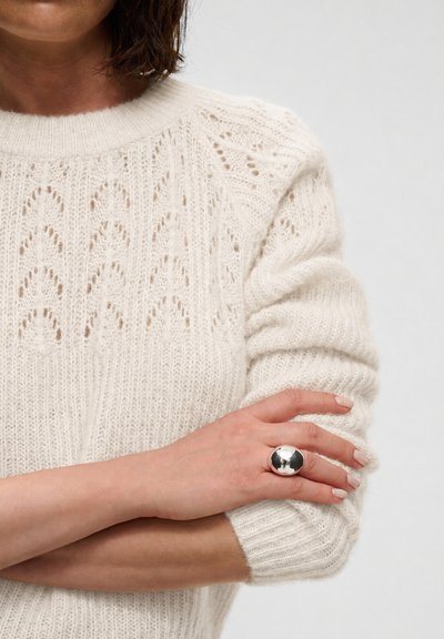 Personne portant un pull en tricot blanc, les bras croisés, montrant une grande bague ronde en argent à une main.