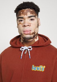 Sudadera con cremallera de color burdeos con un acabado texturizado, que presenta un logo bordado en azul y amarillo y un cordón con un patrón azul y blanco.