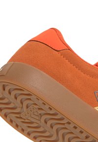 Gros plan du talon d'une basket en daim orange avec une semelle en gomme texturée et le logo Adidas sur le motif de la semelle.