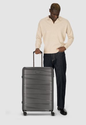 Valigia rigida di colore grigio scuro con motivo a onde testurizzato, manico estensibile e quattro ruote per la mobilità. Chiusura con zip regolabile.