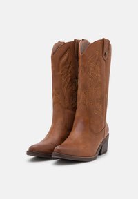 Bottes de cowboy en cuir marron avec un bout pointu, une couture décorative présentant des motifs de feuilles, et un talon empilé.