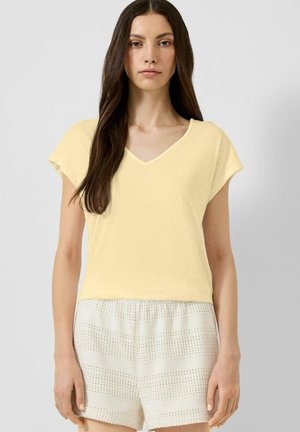Femme aux longs cheveux foncés portant un haut jaune pâle à col en V et manches courtes et un short blanc texturé taille haute.