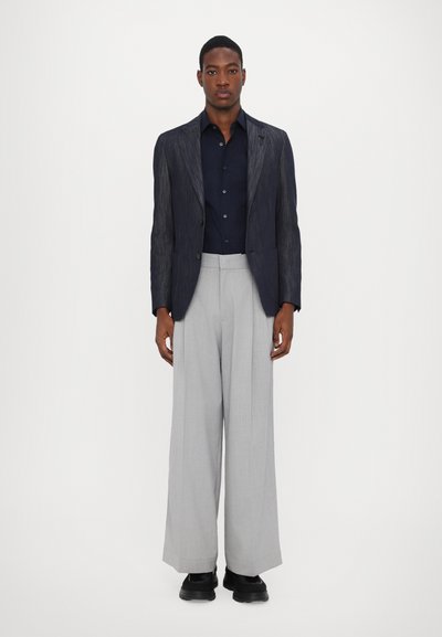 Dunkelblauer Nadelstreifen-Blazer, marineblaues Hemd, hellgraue weite Hose und schwarze Schuhe. Glattes Material und taillierte Passform.