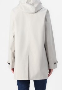 Beige regenjas met capuchon en lange mouwen, met knoopsluitingen aan de mouwen, gedragen over donkerblauwe spijkerbroek, weergegeven vanuit de achterkant.