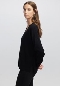 Schwarzer Strickpullover mit V-Ausschnitt, langen Ärmeln und seitlichen Schlitzen. Weiche Textur, lockere Passform und Knopfdetails am Hals.