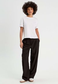 T-shirt bianca in cotone con maniche arrotolate, abbinata a pantaloni neri a vita alta. Il modello indossa sneaker beige, in piedi su uno sfondo neutro.