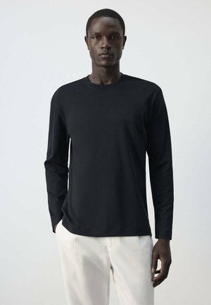 Homme portant une chemise noire uni à manches longues et un pantalon blanc, debout devant un fond clair uni, face à l'avant.