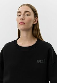 Hanorac negru cu guler rotund cu dungi; prezintă un logo subtil "oui" negru pe piept. Material moale, croială lejeră, design minimalist.