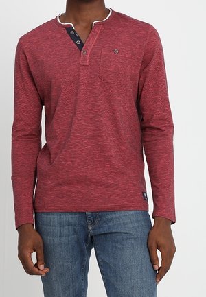 Pitkähihaisen punaisen henley-paidan, jossa on teksturoitu kuvio, valkoinen kaulus, kolme nappia edessä ja rintatasku, yhdistettynä vaaleansinihin farkkuihin.