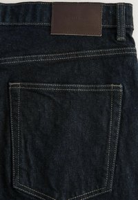 Jeans de mezclilla oscura con una parche de cuero marrón en la cintura y costuras en contraste. Incluye un solo bolsillo trasero con un acabado limpio.