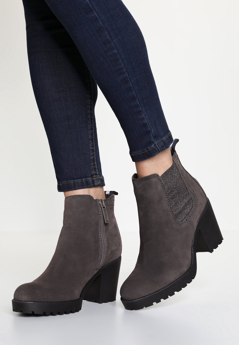 Bottes en daim gris foncé montant jusqu'à la cheville, avec talons épais et fermeture éclair sur le côté, portées avec un jean ajusté bleu foncé.