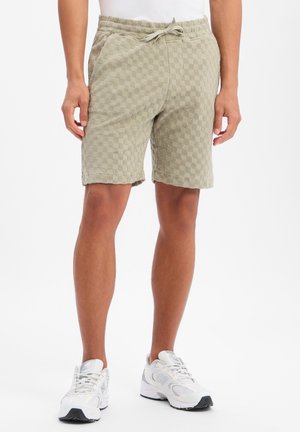 Homme portant un short beige texturé avec cordon de serrage et des baskets blanches, debout devant un fond blanc uni.
