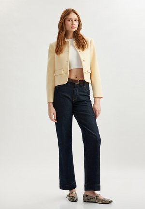 Cremfarbener Blazer mit goldenen Knöpfen über einem weißen, knappen Top, kombiniert mit dunkelblauen, geraden Jeans und Schlangenleder-Ballerinas.